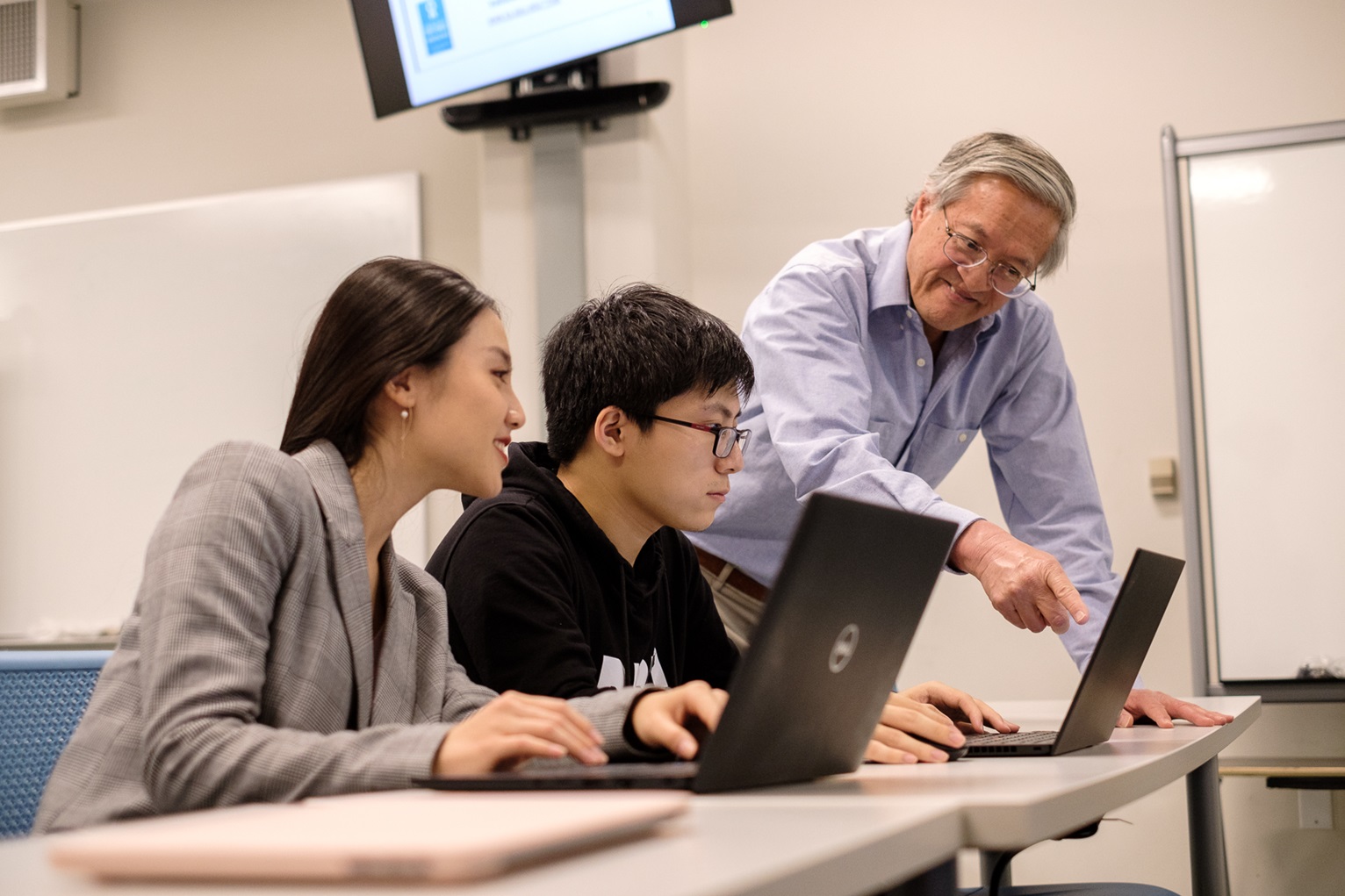 Two students sit at a desk in front of a computer with a professor teaching them at SJSU.