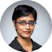 Karthika Sasikumar A person with short black hair and blue glasses is wearing a black shirt and gold jewelry against a plain backdrop.