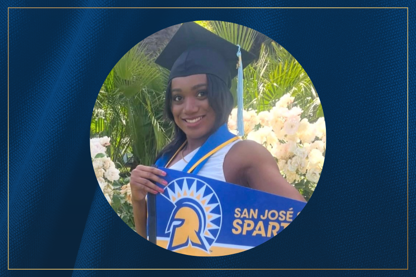 A proud graduate in a cap and gown poses with a blue and gold sign celebrating their achievement at San Jose State University.