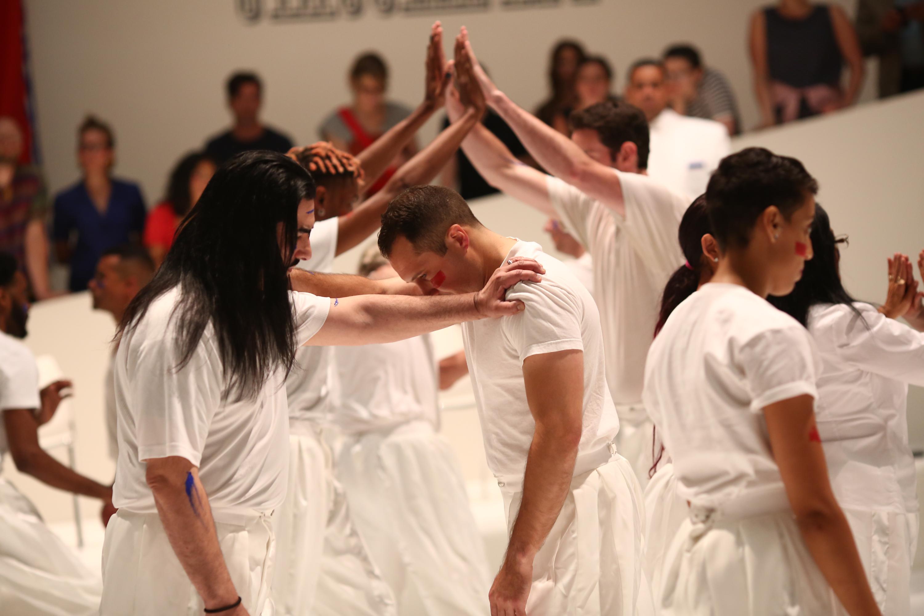 Image caption: Primitive Games - performance, 1 hr. at Guggenheim Museum, New York, NY, 6/21/18. Photo by Paula Court performance still of figures wearing white clothing and joining hands.  The two figures in the foreground rest their hands on each other shoulders